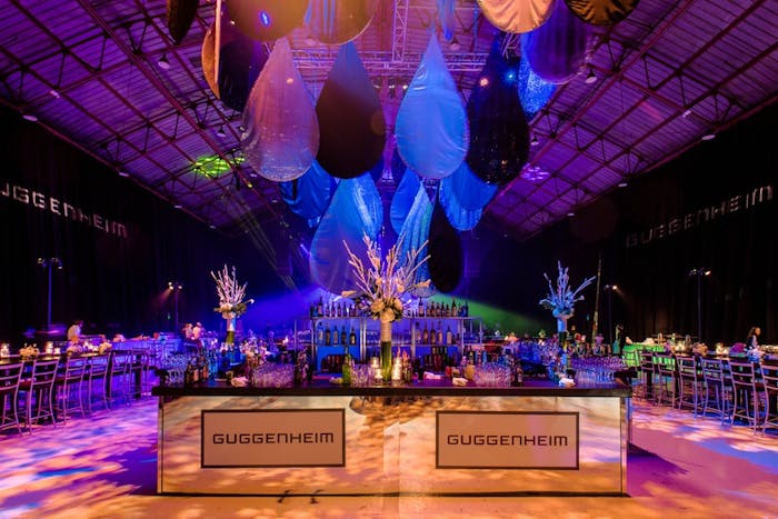 a mirrored bar in the foreground with blue lighting and a funky chandelier hanging above. Tables and chairs are behind the bar.