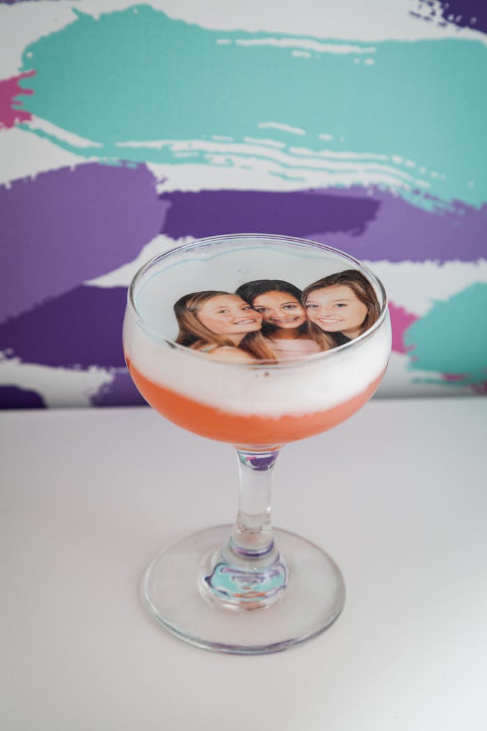 a pink drink in a sidecar glass with foam on top and a photo of three girls laid on top 