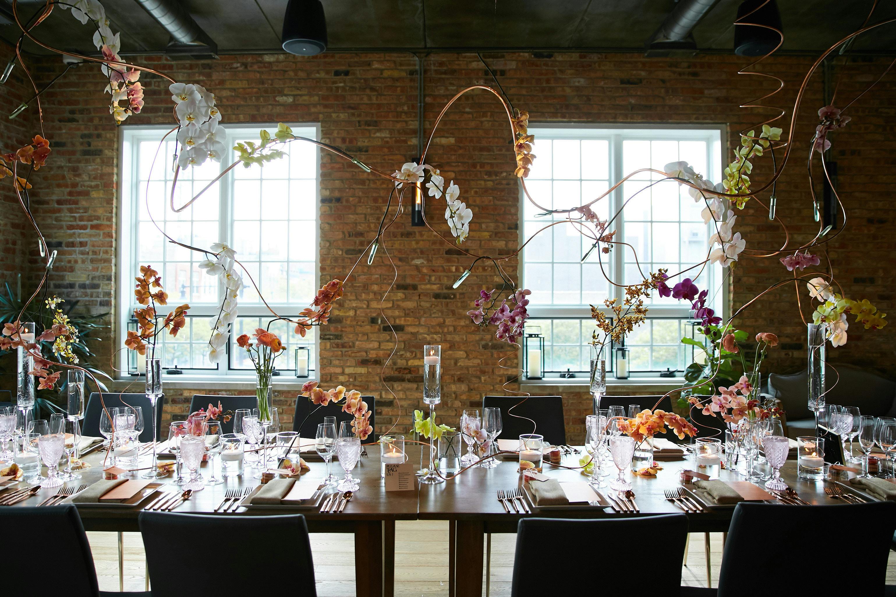 a table in the foreground holds geometric and sparse floral arrangements that extent a few feet into the air. The background holds a brick wall with 2 windows that showcase natural light.