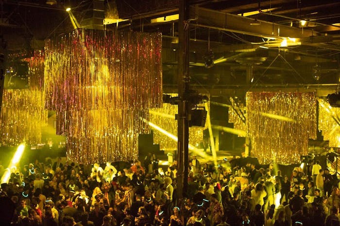 large space with gold tinsel hanging from ceiling. People dance underneath the yellow dance lighting while yellow lazer lights point to the dance floor.