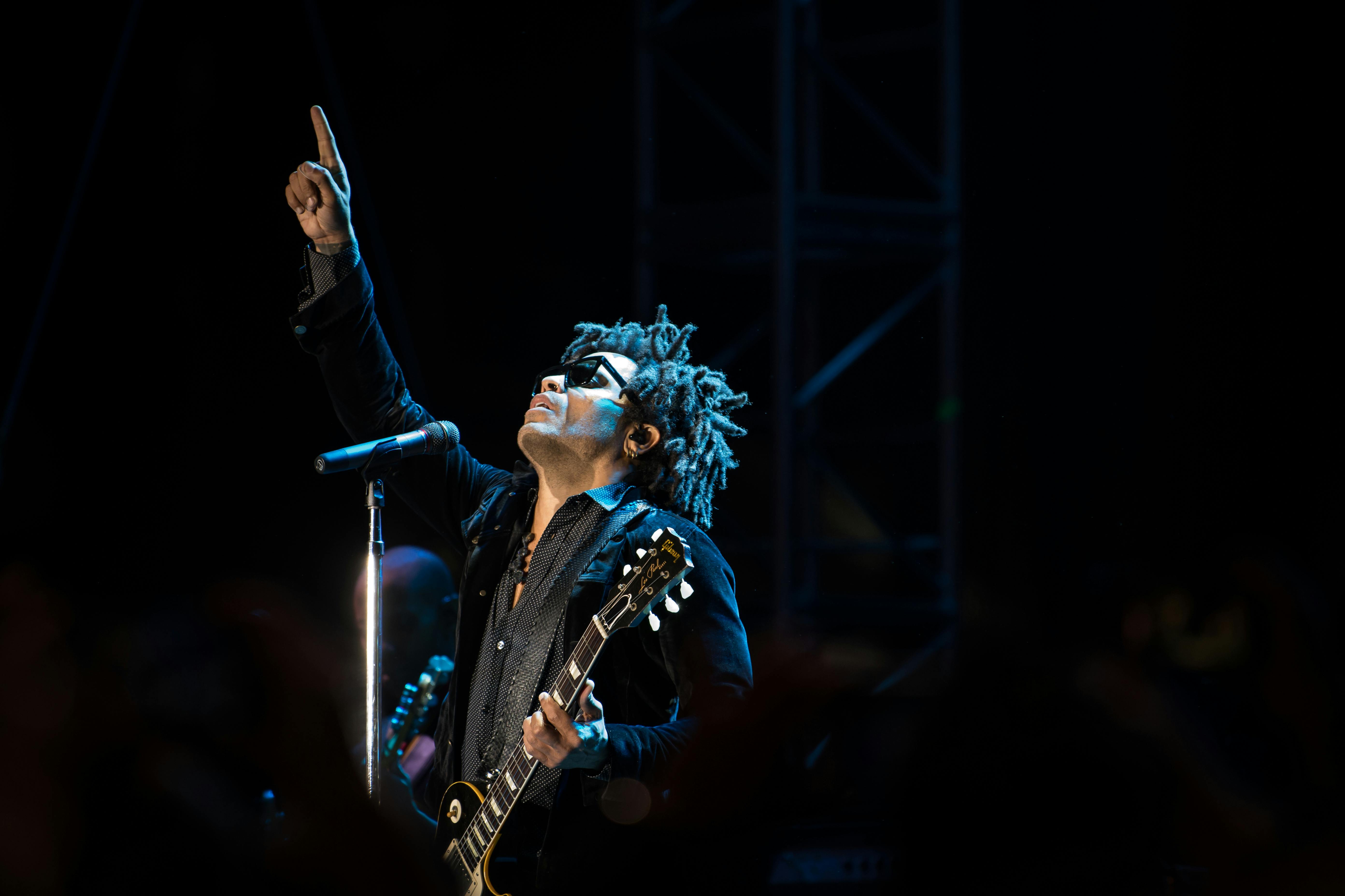a singer on stage points to the sky while holding his guitar.