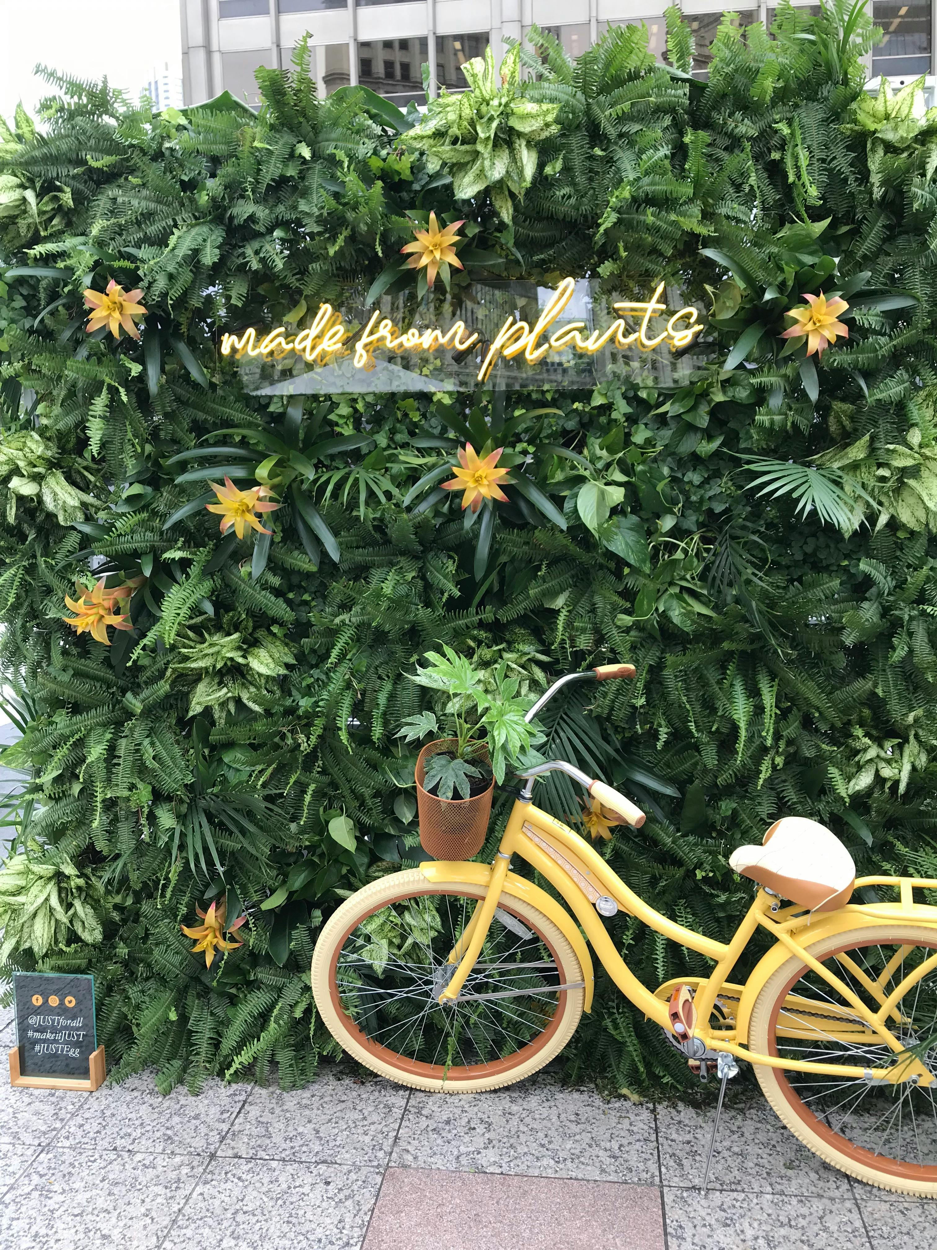 a yellow vintage bike against a huge green bush. Yellow flowers and a yellow neon sign that says "made from plants" is on the bush. 