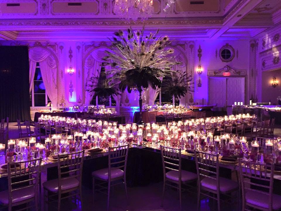 Purple room with tables arranged in x and large peacock feathers in the middle. The tables have candles throughout making them glow.