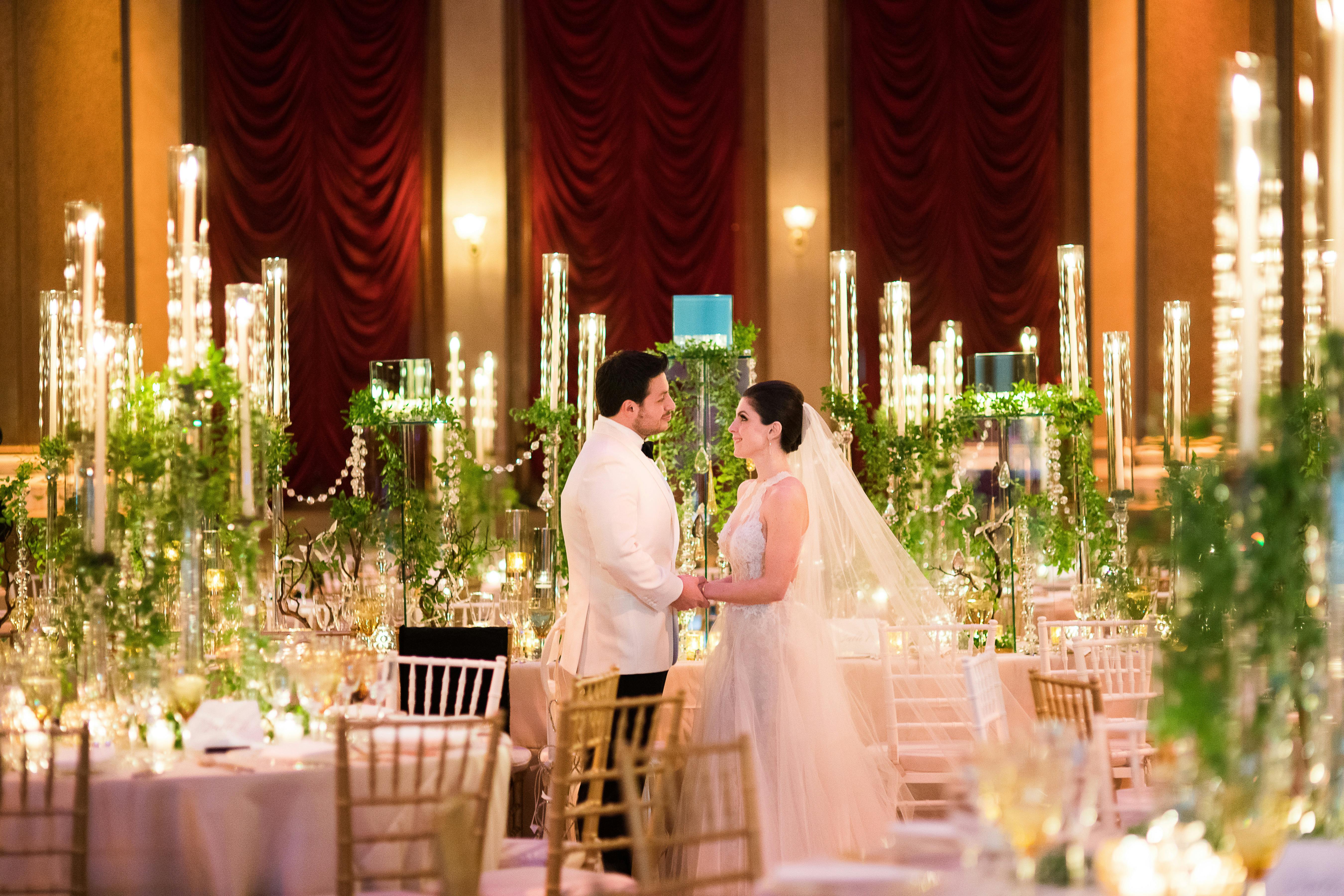 Husband and Wife stand in between tables with candelabras draped in greenery.