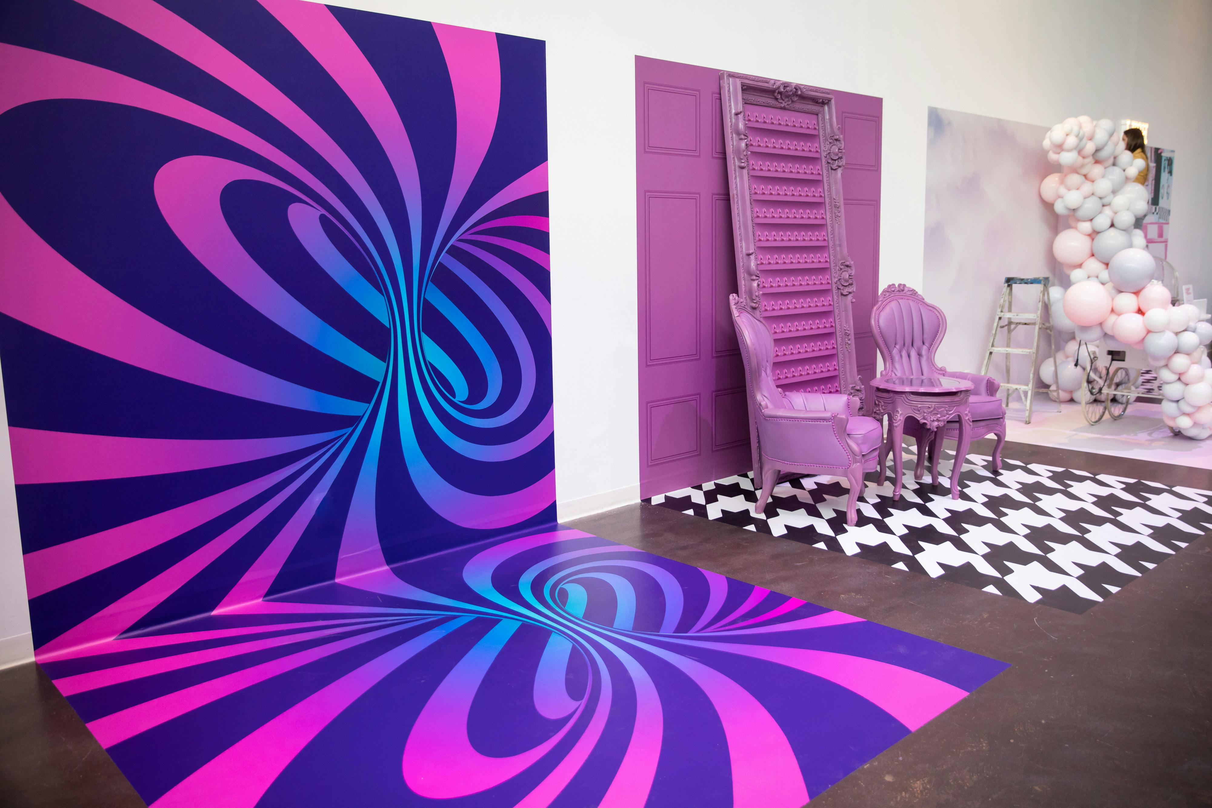 various instagram backdrops along a white wall. The first is a spiral effect that is pink, purple, and blue. the second is a pink barn door with two victorian style chairs and the third is a cloud of balloons. 