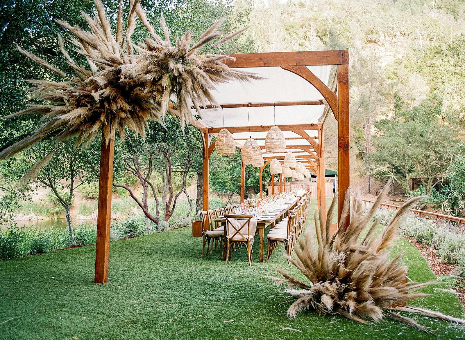 A cherry stained pergola with pampas grass accents. Underneath is a long table with white cushioned chairs on either side.