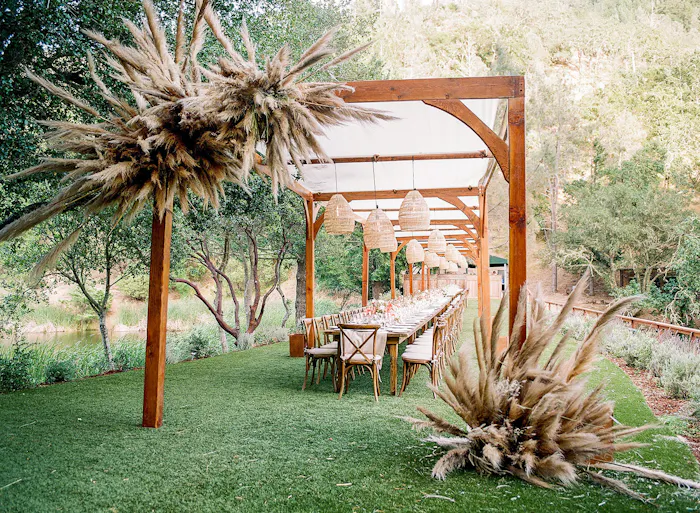 A cherry stained pergola with pampas grass accents. Underneath is a long table with white cushioned chairs on either side.