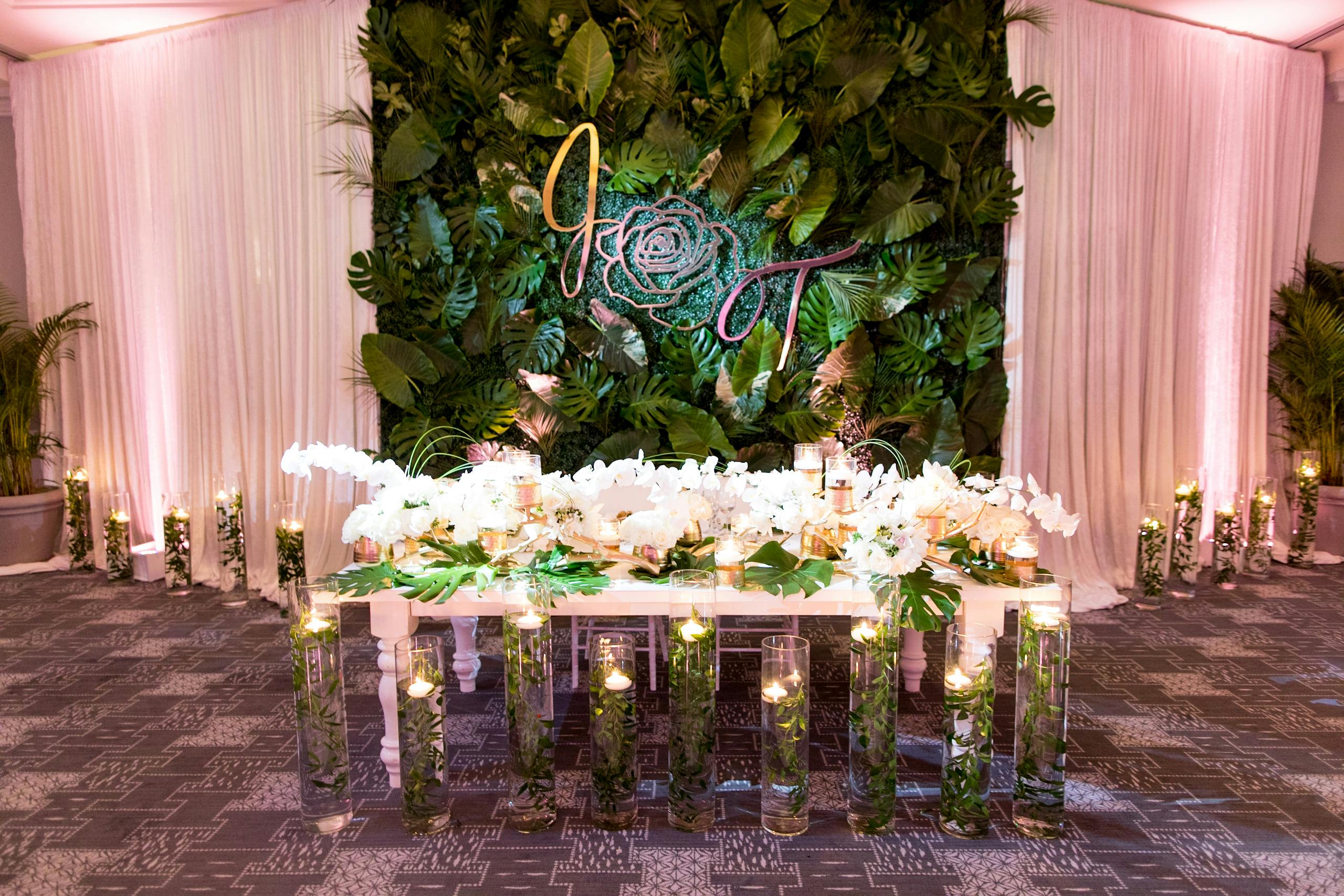 Candlelit ballroom shot of table with white orchids against backdrop of pale pink curtain and tropical leaves. 