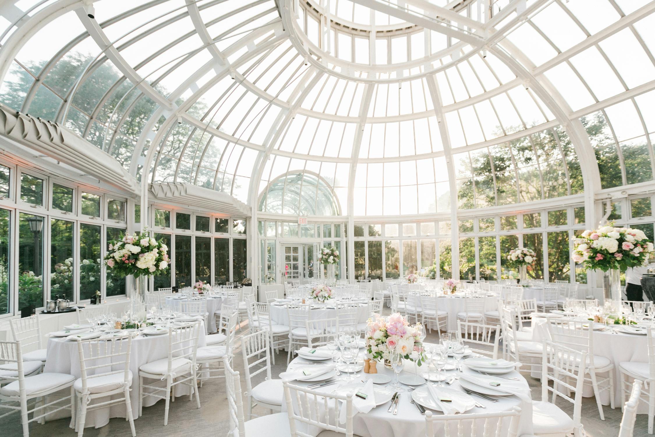 A large paned glass structure with all white furniture and short floral arrangements. 