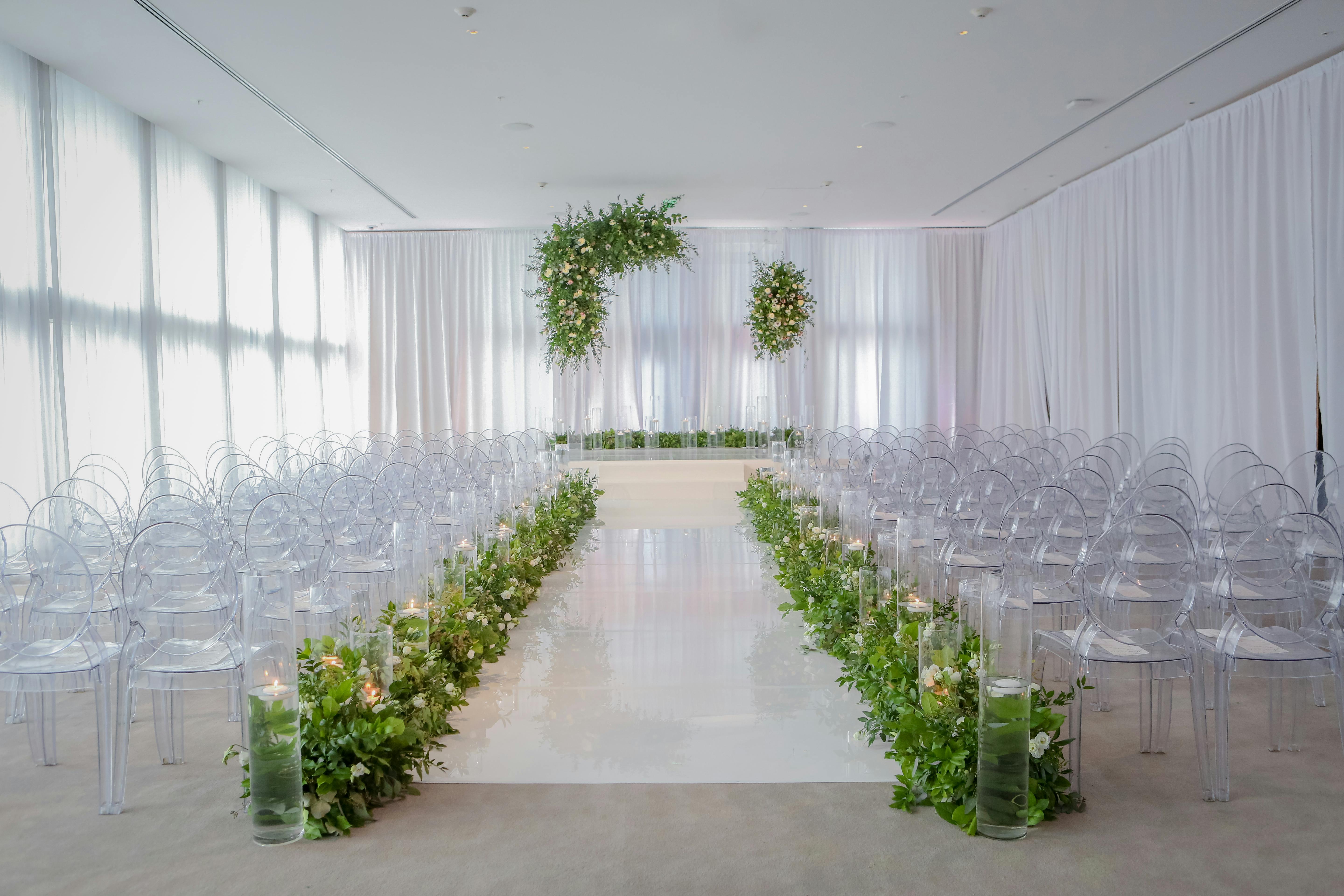 Clear chairs on either side of the aisle and an all white color palette. Greenery lines the aisle as well as drapes over the altar.